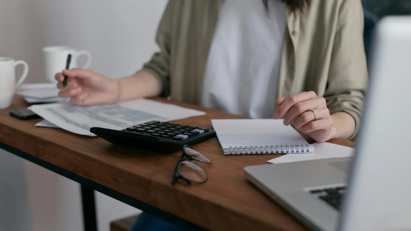 A woman manages finances at home, using a laptop and calculator on a wooden desk., tags: für und vermieter ab - pexel
