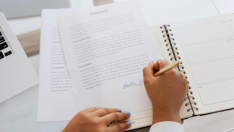 Close-up of hands signing a contract on a desk with office supplies, symbolizing legal agreements., tags: ausland - pexel